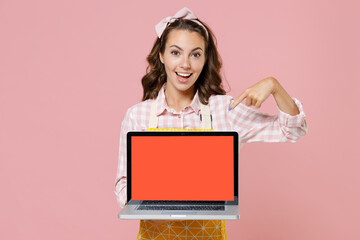 Excited young woman housewife in yellow apron pointing index finger on laptop computer with blank...