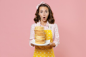 Shocked amazed young brunette woman housewife 20s wearing yellow apron hold plate with pancakes while doing housework isolated on pastel pink colour background studio portrait. Housekeeping concept.