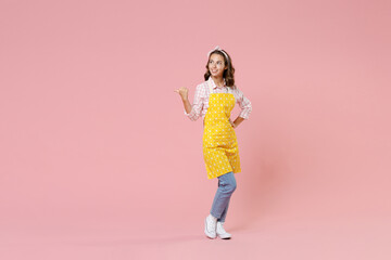 Fototapeta na wymiar Full length portrait of smiling young woman housewife wearing yellow apron pointing thumb aside on mock up copy space doing housework isolated on pastel pink background studio. Housekeeping concept.