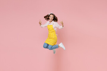 Full length portrait of cheerful young brunette woman housewife 20s in yellow apron jumping showing thumbs up doing housework isolated on pastel pink colour background studio. Housekeeping concept.