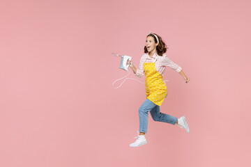 Fototapeta na wymiar Full length portrait of excited young brunette woman housewife 20s in yellow apron jumping holding mixer while doing housework isolated on pastel pink colour background studio. Housekeeping concept.