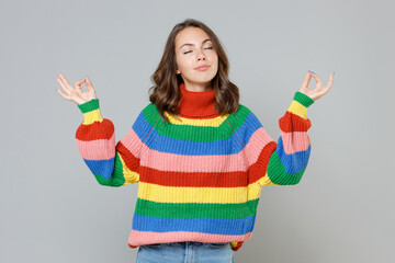 Young brunette woman 20s in casual colorful knitted sweater hold hands in yoga gesture, relaxing meditating, trying to calm down keeping eyes closed isolated on grey colour background studio portrait.