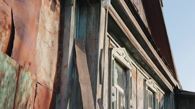 An old wooden small house with peeling paint on the walls with brown loft on sunny day. An outbuilding is paneled with iron next to decrepit Russian hut.