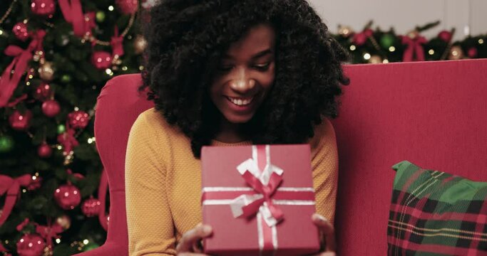 Beautiful african american woman opening Christmas present while sitting at home on red sofa. Happy woman reacts emotionally and surprised to gift .