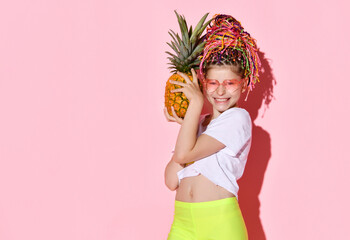Teenage female with african pigtails, in sunglasses. She is holding pineapple and smiling, posing on pink background. Close up
