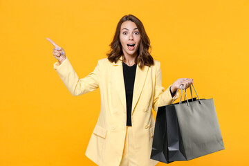 Shocked young woman in suit jacket hold package bags with purchases after shopping point index finger aside up on mock up copy space isolated on yellow background studio portrait. Black friday sale.