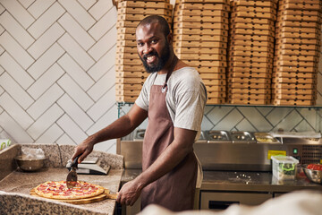 Handsome Afro American man cutting pepperoni pizza in pizzeria - obrazy, fototapety, plakaty