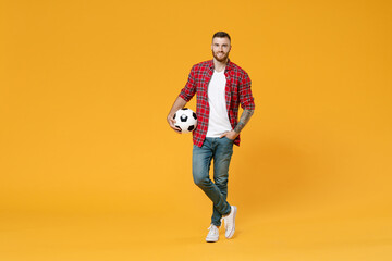 Full length portrait of smiling young man football fan in red shirt cheer up support favorite team with soccer ball looking camera isolated on yellow background studio. People sport leisure concept.