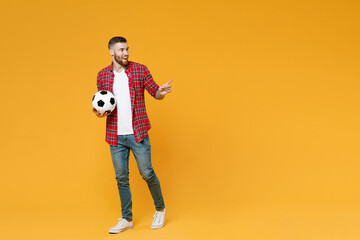 Fototapeta premium Full length portrait funny young man football fan in red shirt cheer up support favorite team with soccer ball pointing index finger aside isolated on yellow background. People sport leisure concept.