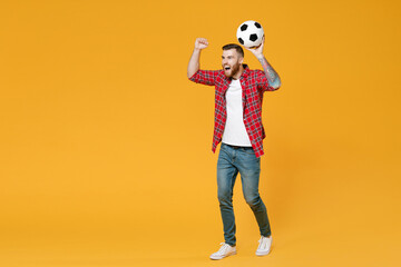 Full length portrait screaming young man football fan in red shirt cheer up support favorite team with soccer ball clenching fists isolated on yellow background studio. People sport leisure concept.