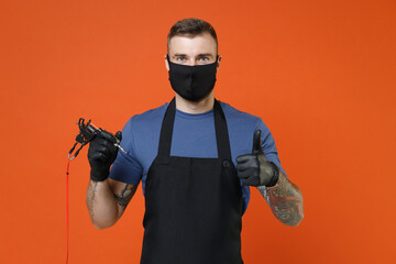 Young professional tattooer master artist tattooed man in t-shirt apron face mask hold machine black ink in jar equipment for making tattoo art on body showing thumb up isolated on brown background.