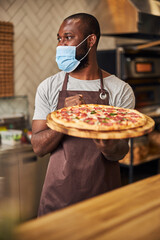 Afro American man in protective face mask holding pepperoni pizza