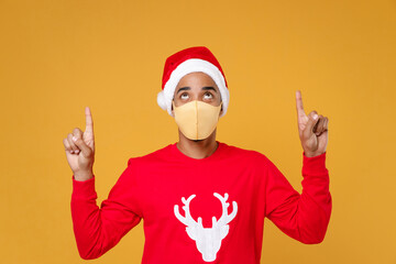 Fototapeta na wymiar Young Santa african american man in Christmas hat face mask to safe from coronavirus virus covid-19 pointing index fingers up isolated on yellow background. Happy New Year celebration holiday concept.