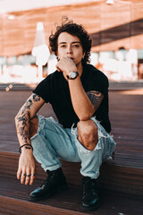 Hombre joven adolescente con tatuajes modelo en una escalera con camiseta negra  y pantalones...