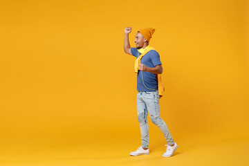 Fototapeta premium Full length of joyful young african american man 20s wearing basic blue t-shirt hat standing clenching fists doing winner gesture looking aside isolated on bright yellow background, studio portrait.