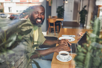 Cheerful Afro American man behind glass working on laptop