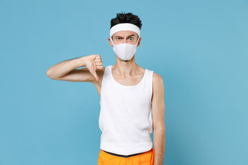 Fototapeta na wymiar Young sporty man with skinny body sportsman in headband shirt face mask to safe from coronavirus virus covid-19 showing thumb down isolated on blue background. Workout gym sport motivation concept.