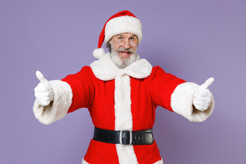 Fototapeta na wymiar Excited elderly gray-haired Santa Claus man in Christmas hat red suit coat glasses showing thumbs up isolated on violet purple background studio. Happy New Year celebration merry holiday concept.