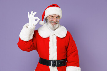 Fototapeta na wymiar Cheerful elderly gray-haired Santa Claus man in Christmas hat red suit coat glasses showing OK gesture isolated on violet purple background studio. Happy New Year celebration merry holiday concept.