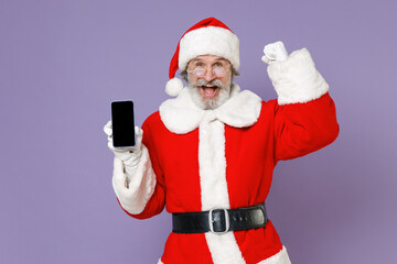 Fototapeta na wymiar Joyful Santa Claus man in Christmas hat red coat glasses hold mobile phone with blank empty screen doing winner gesture isolated on violet background. Happy New Year celebration merry holiday concept.