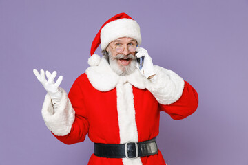 Excited Santa Claus man in Christmas hat red suit coat white gloves glasses talking on mobile phone spreading hands isolated on violet background. Happy New Year celebration merry holiday concept.