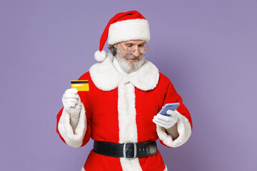 Elderly Santa Claus man in Christmas hat red suit coat white gloves glasses using mobile phone hold credit bank card isolated on violet background. Happy New Year celebration merry holiday concept.
