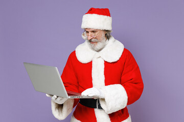 Naklejka premium Elderly gray-haired Santa Claus man in Christmas hat red suit coat white gloves glasses working on laptop pc computer isolated on violet background. Happy New Year celebration merry holiday concept.