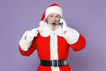 Fototapeta na wymiar Shocked Santa Claus man in Christmas hat red suit coat white gloves glasses talking point index finger on mobile phone isolated on violet background. Happy New Year celebration merry holiday concept.