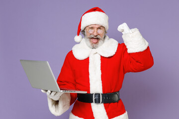Blinking Santa Claus man in Christmas hat red coat gloves glasses working on laptop pc computer doing winner gesture isolated on violet background. Happy New Year celebration merry holiday concept.