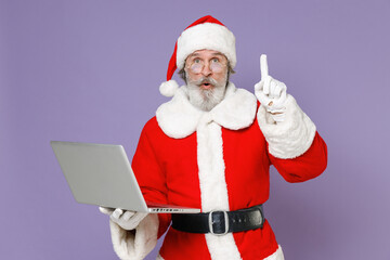 Fototapeta na wymiar Amazed Santa Claus man in Christmas hat red coat working on laptop pc computer hold index finger up with great new idea isolated on violet background. Happy New Year celebration merry holiday concept.