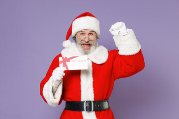 Fototapeta na wymiar Joyful elderly Santa Claus man in Christmas hat red suit coat gloves glasses hold gift certificate doing winner gesture isolated on violet background. Happy New Year celebration merry holiday concept.
