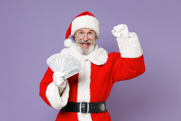 Joyful Santa Claus man in Christmas hat red suit coat glasses hold fan cash money dollar banknotes doing winner gesture isolated on violet background. Happy New Year celebration merry holiday concept.