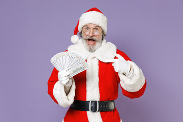 Fototapeta na wymiar Amazed Santa Claus man in Christmas hat red suit coat glasses point index finger on fan of cash money dollar banknotes isolated on violet background. Happy New Year celebration merry holiday concept.