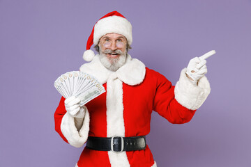 Fototapeta na wymiar Funny Santa Claus man in Christmas hat red suit coat hold fan of cash money dollar banknotes point index finger aside isolated on violet background. Happy New Year celebration merry holiday concept.