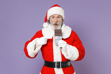 Fototapeta na wymiar Shocked Santa Claus man in Christmas hat red suit coat white gloves glasses point index finger on chocolate bar isolated on violet background studio. Happy New Year celebration merry holiday concept.