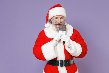 Fototapeta na wymiar Cheerful gray-haired Santa Claus man in Christmas hat red suit coat white gloves glasses biting chocolate bar isolated on violet background studio. Happy New Year celebration merry holiday concept.
