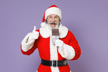 Fototapeta na wymiar Excited Santa Claus man in Christmas hat red suit coat white gloves glasses hold chocolate bar showing thumb up isolated on violet background studio. Happy New Year celebration merry holiday concept.