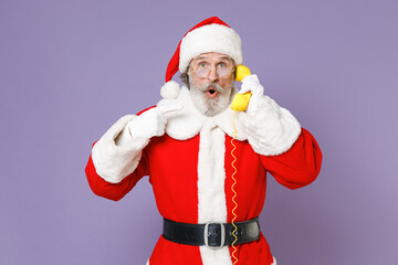 Fototapeta na wymiar Shocked Santa Claus man in Christmas hat red suit coat gloves glasses talking pointing index finger on telephone isolated on violet background studio. Happy New Year celebration merry holiday concept.