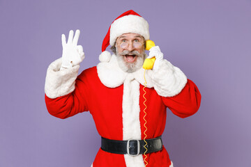 Excited Santa Claus man in Christmas hat red suit coat gloves glasses talking on telephone showing OK gesture isolated on violet background studio. Happy New Year celebration merry holiday concept.