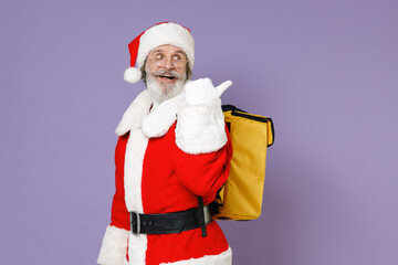 Side view funny Santa Claus man in Christmas hat red suit coat glasses thermal food bag backpack pointing thumb aside isolated on violet background. Happy New Year celebration merry holiday concept.