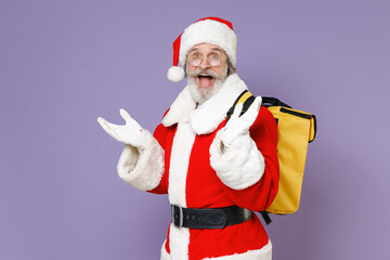 Fototapeta na wymiar Side view of surprised Santa Claus man in Christmas hat red suit coat glasses thermal food bag backpack spreading hands isolated on violet background. Happy New Year celebration merry holiday concept.