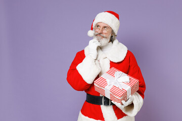 Pensive Santa Claus man in Christmas hat red suit coat hold present box with gift ribbon bow put hand prop up on chin isolated on violet background. Happy New Year celebration merry holiday concept.