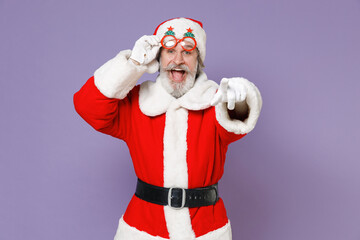 Fototapeta na wymiar Excited elderly Santa Claus man in Christmas hat red suit coat white gloves glasses point index finger on camera isolated on violet background studio. Happy New Year celebration merry holiday concept.