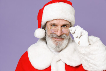 Smiling elderly gray-haired Santa Claus man wearing Christmas hat red suit coat glasses looking camera isolated on violet purple background studio. Happy New Year celebration merry holiday concept.