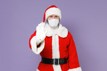 Santa Claus man in Christmas hat suit coat face mask to safe from coronavirus virus covid-19 showing stop gesture with palm isolated on violet background. New Year celebration merry holiday concept.