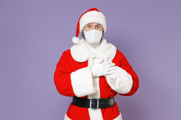 Fototapeta na wymiar Santa Claus man in Christmas hat red coat face mask to safe from coronavirus virus covid-19 hold hands folded on heart isolated on violet background. Happy New Year celebration merry holiday concept.