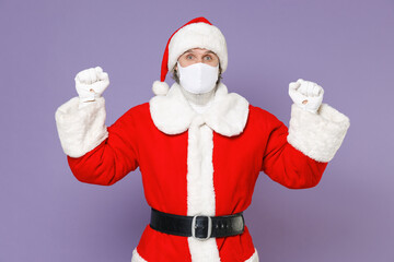 Fototapeta na wymiar Santa Claus man in Christmas hat red coat suit face mask to safe from coronavirus virus covid-19 doing winner gesture isolated on violet background. Happy New Year celebration merry holiday concept.