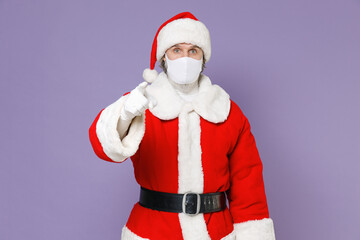 Fototapeta na wymiar Santa Claus man in Christmas hat red coat mask to safe from coronavirus virus covid-19 pointing index finger on camera isolated on violet background. Happy New Year celebration merry holiday concept.