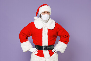 Fototapeta na wymiar Santa Claus man in Christmas hat red coat face mask safe from coronavirus virus covid-19 standing with arms akimbo on waist isolated on violet background. New Year celebration merry holiday concept.