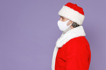 Side view of Santa Claus man in Christmas hat red suit face mask safe from coronavirus virus covid-19 during quarantine isolated on violet background. Happy New Year celebration merry holiday concept.